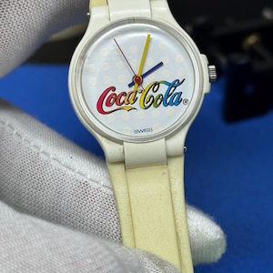 May include: A vintage white wristwatch with a white dial featuring the Coca-Cola logo in red, yellow, and blue. The watch has a white band and a Swiss movement. The watch hands are yellow, blue, and red.