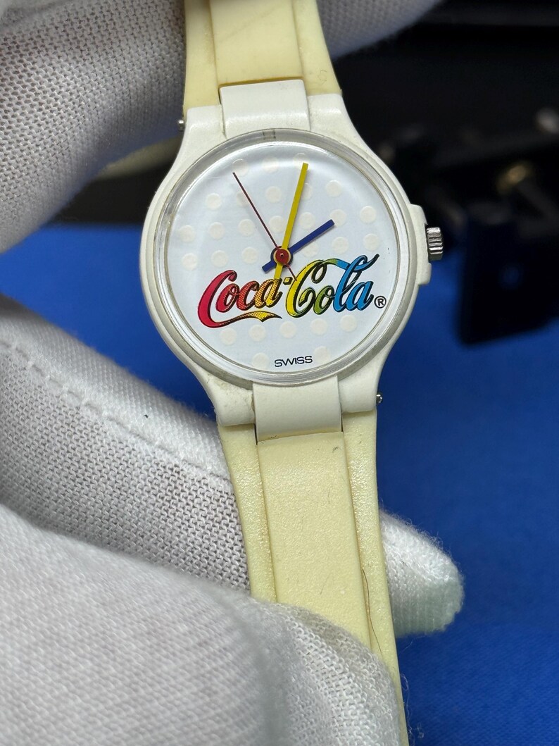 May include: A vintage white wristwatch with a white dial featuring the Coca-Cola logo in red, yellow, and blue. The watch has a white band and a Swiss movement. The watch hands are yellow, blue, and red.