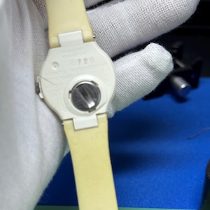 May include: A close-up of the back of a cream-colored wristwatch with a matching strap. The watch face is white with a silver battery compartment. The strap is a light yellow color. The watch is held by a gloved hand.