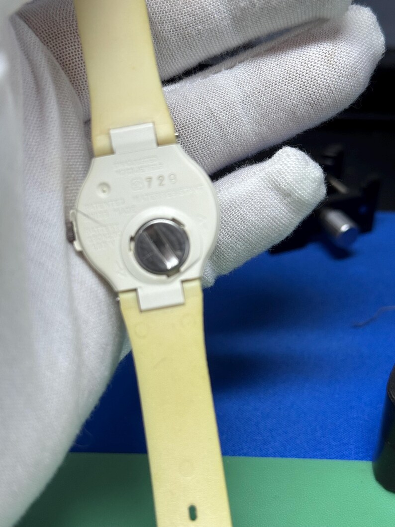 May include: A close-up of the back of a cream-colored wristwatch with a matching strap. The watch face is white with a silver battery compartment. The strap is a light yellow color. The watch is held by a gloved hand.
