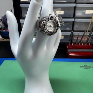 Op de afbeelding: Een zilverkleurige ringhorloge met een witte wijzerplaat en zwarte cijfers. Het horloge heeft een sierlijke, sierlijke rand en wordt gepresenteerd op een witte handvormige display. Een heldere, rechthoekige steen is naast het horloge geplaatst.