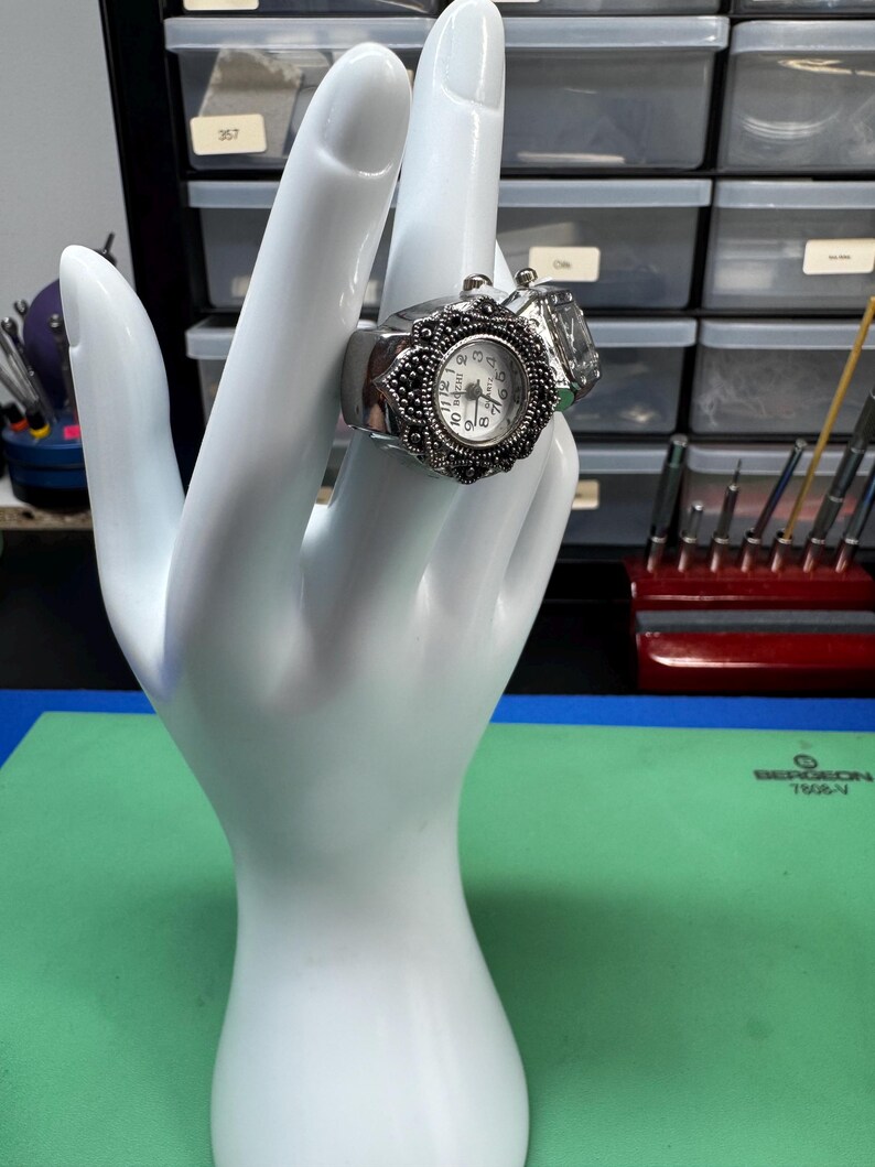 Op de afbeelding: Een zilverkleurige ringhorloge met een witte wijzerplaat en zwarte cijfers. Het horloge heeft een sierlijke, sierlijke rand en wordt gepresenteerd op een witte handvormige display. Een heldere, rechthoekige steen is naast het horloge geplaatst.
