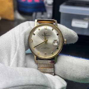 Pode incluir: Um relógio de pulso dourado com um mostrador branco e ponteiros pretos. O relógio tem uma pulseira dourada e uma janela de data na posição das 3 horas. O mostrador do relógio diz "Roy Richard" e "17 Jewels Shockresistant".