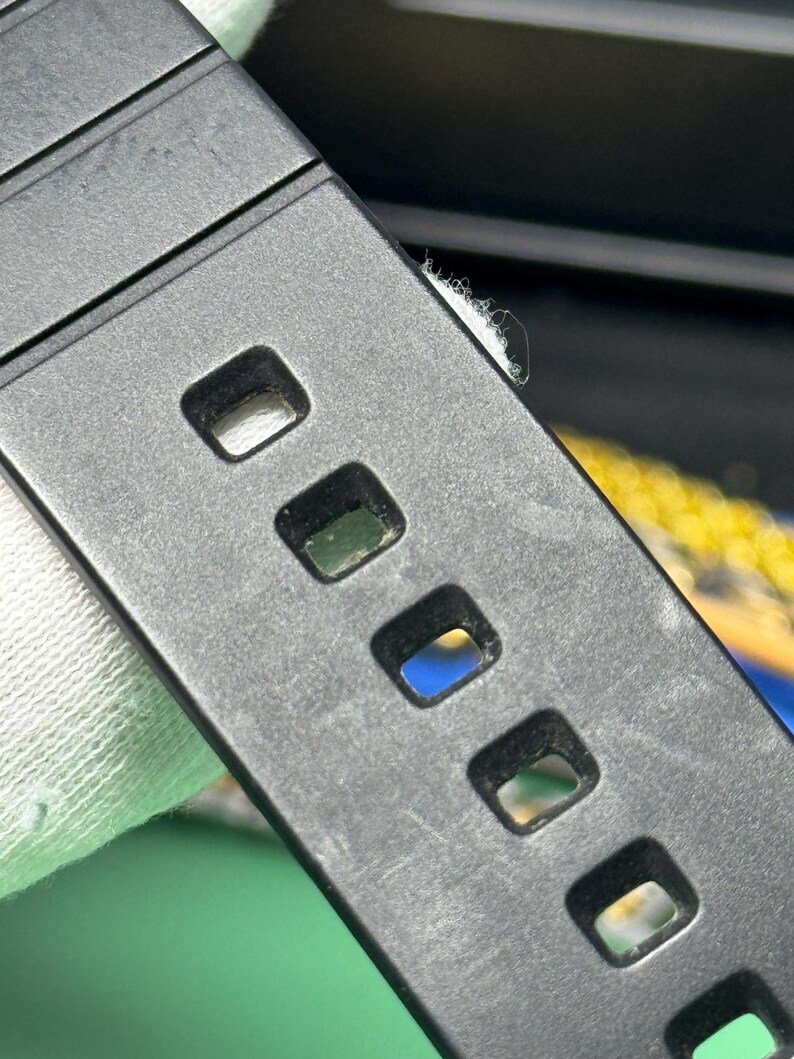 Peut inclure: Gros plan sur un bracelet de montre noir avec des trous rectangulaires. Le bracelet est fait d'un mat&eacute;riau textur&eacute; et est inclin&eacute; en diagonale dans le cadre. L'arri&egrave;re-plan est flou, avec des indices d'autres couleurs.