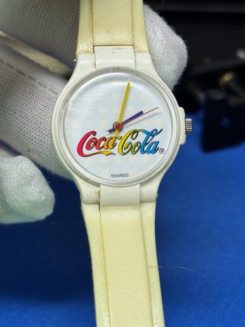 May include: A vintage white wristwatch featuring the Coca-Cola logo in red, yellow, and blue. The watch has a white dial with a dotted pattern and yellow, blue, and red hands. The watch band is a faded cream color, and the word "SWISS" is printed on the watch face.