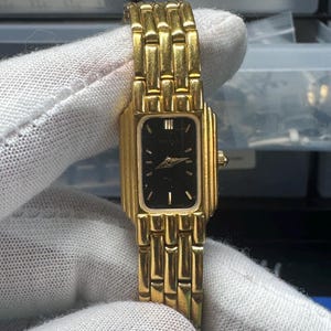May include: A gold-toned wristwatch with a rectangular black face. The watch features a gold band with a series of rectangular links. The watch face has gold hands and hour markers. The watch is held by a gloved hand.