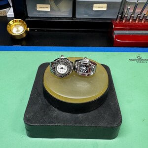 Op de afbeelding: Twee ringen met horlogezichten, &eacute;&eacute;n met een zilveren filigraanontwerp en de andere met een rechthoekig gezicht en een rood midden. Ze staan op een geel en zwart statief. Op de achtergrond is een kleine goudkleurige klok te zien.