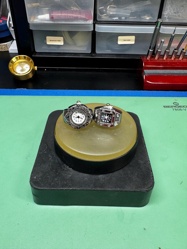 Op de afbeelding: Twee ringen met horlogezichten, &eacute;&eacute;n met een zilveren filigraanontwerp en de andere met een rechthoekig gezicht en een rood midden. Ze staan op een geel en zwart statief. Op de achtergrond is een kleine goudkleurige klok te zien.