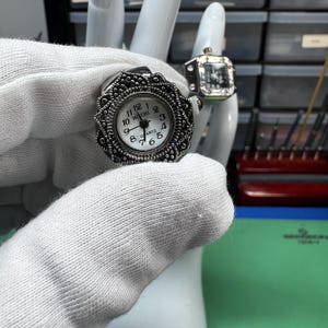 Op de afbeelding: Er worden twee ringhorloges getoond. De eerste is een zilverkleurig ringhorloge met een ronde wijzerplaat en een decoratieve rand. De wijzerplaat is wit met zwarte cijfers en wijzers. Het tweede ringhorloge heeft een vierkante wijzerplaat.