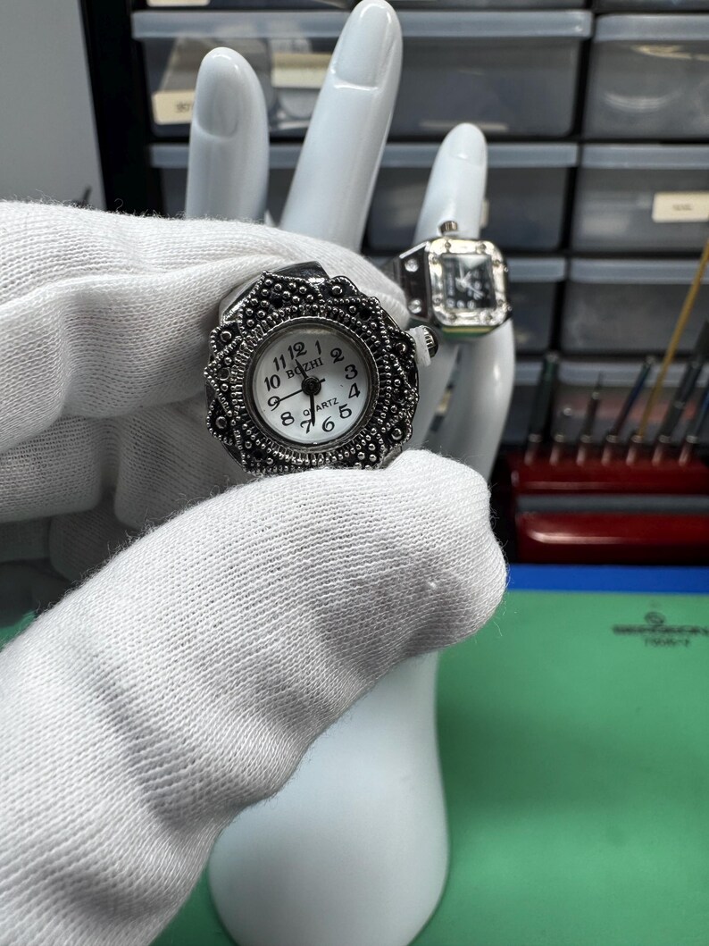 Op de afbeelding: Er worden twee ringhorloges getoond. De eerste is een zilverkleurig ringhorloge met een ronde wijzerplaat en een decoratieve rand. De wijzerplaat is wit met zwarte cijfers en wijzers. Het tweede ringhorloge heeft een vierkante wijzerplaat.