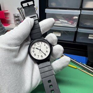 Peut inclure: Montre Casio noire avec un cadran blanc et des chiffres noirs. La montre a un bracelet noir et une lunette noire. La montre est tenue par une main gant&eacute;e.