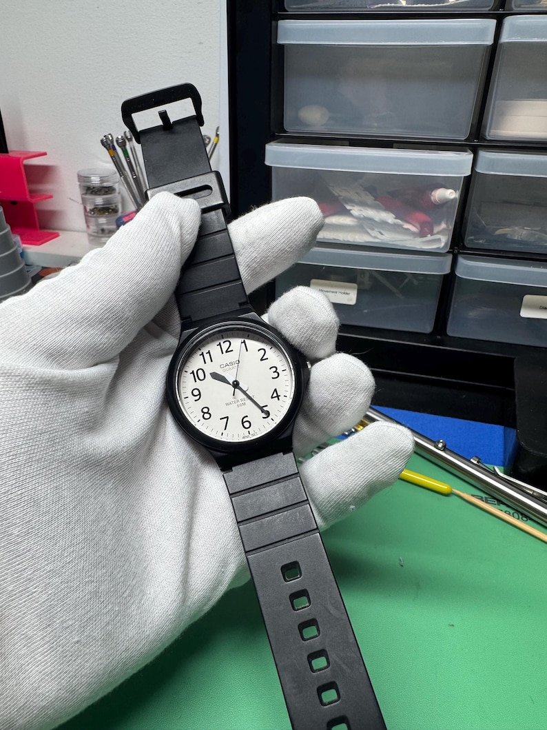 Peut inclure: Montre Casio noire avec un cadran blanc et des chiffres noirs. La montre a un bracelet noir et une lunette noire. La montre est tenue par une main gant&eacute;e.