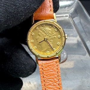May include: A gold-toned wristwatch featuring a coin-style dial with a profile of Liberty and the word LIBERTY inscribed at the top. The watch has a textured gold bezel, thin black hands, and a light brown, textured leather strap with visible stitching.