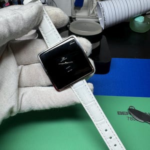 May include: A white leather watch strap with a square black face watch. The watch face has silver hands and a silver border. The watch face says "QUARTZ" in silver lettering.
