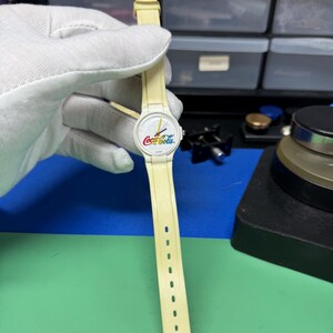 May include: A vintage Coca-Cola wristwatch with a cream-colored band and face. The watch face features the Coca-Cola logo in red, white, and yellow. The watch band has a series of holes for adjustment.