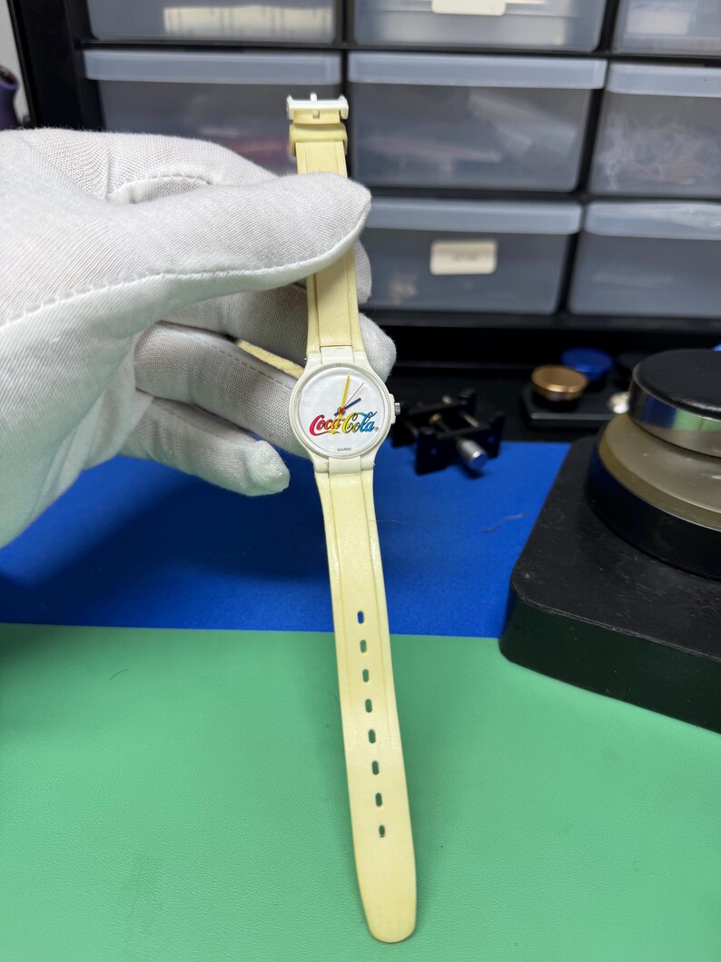May include: A vintage Coca-Cola wristwatch with a cream-colored band and face. The watch face features the Coca-Cola logo in red, white, and yellow. The watch band has a series of holes for adjustment.