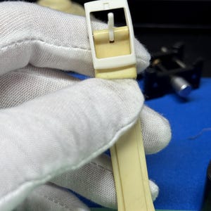May include: Close-up of a cream-colored watch strap and buckle. The buckle is rectangular with a white frame and a cream-colored pin. The watch strap is a matching cream color. The watch face is white with yellow and black accents.