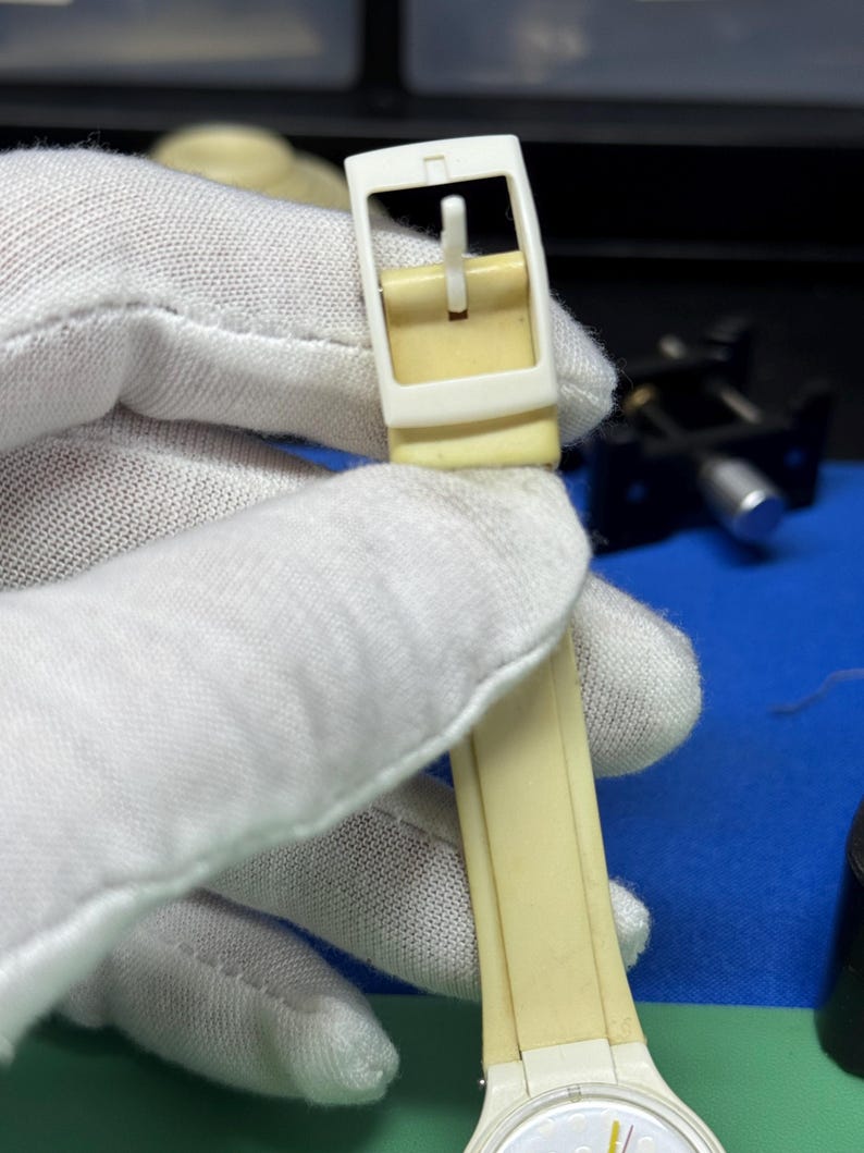 May include: Close-up of a cream-colored watch strap and buckle. The buckle is rectangular with a white frame and a cream-colored pin. The watch strap is a matching cream color. The watch face is white with yellow and black accents.
