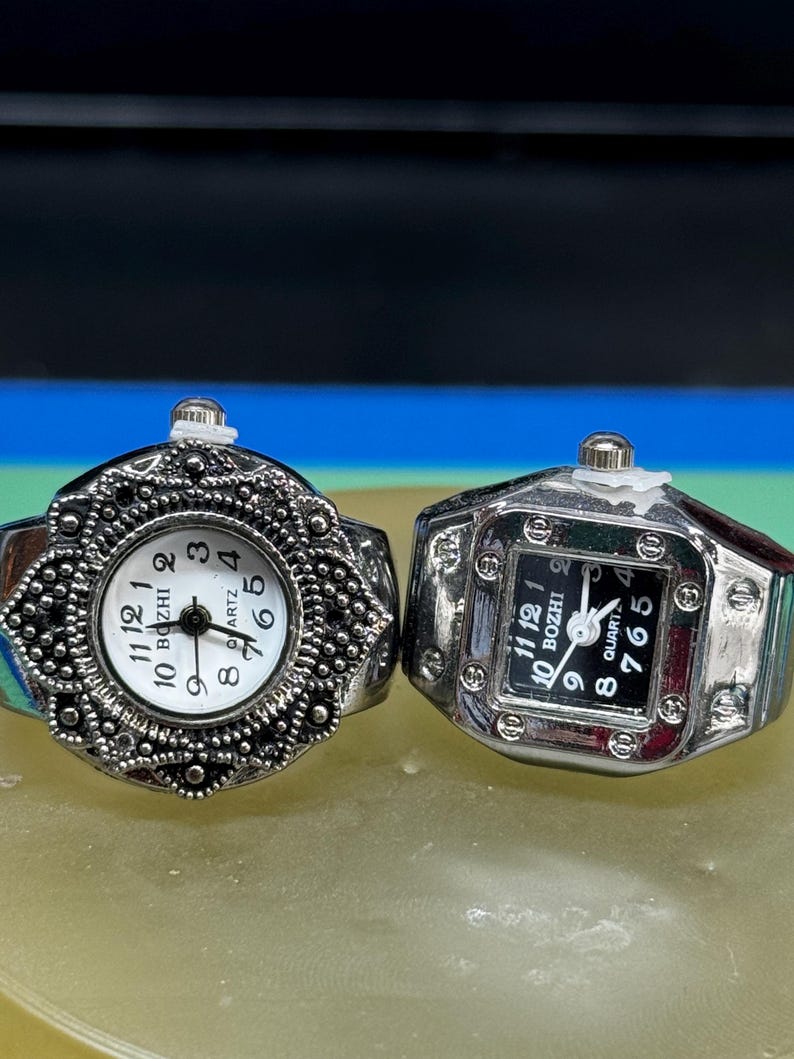 Op de afbeelding: Twee zilverkleurige ringhorloges. E&eacute;n heeft een ronde witte wijzerplaat met zwarte cijfers en de woorden "BOZHI" en "QUARTZ". De andere heeft een vierkante zwarte wijzerplaat met witte cijfers en dezelfde tekst. Beide hebben een decoratief ontwerp.