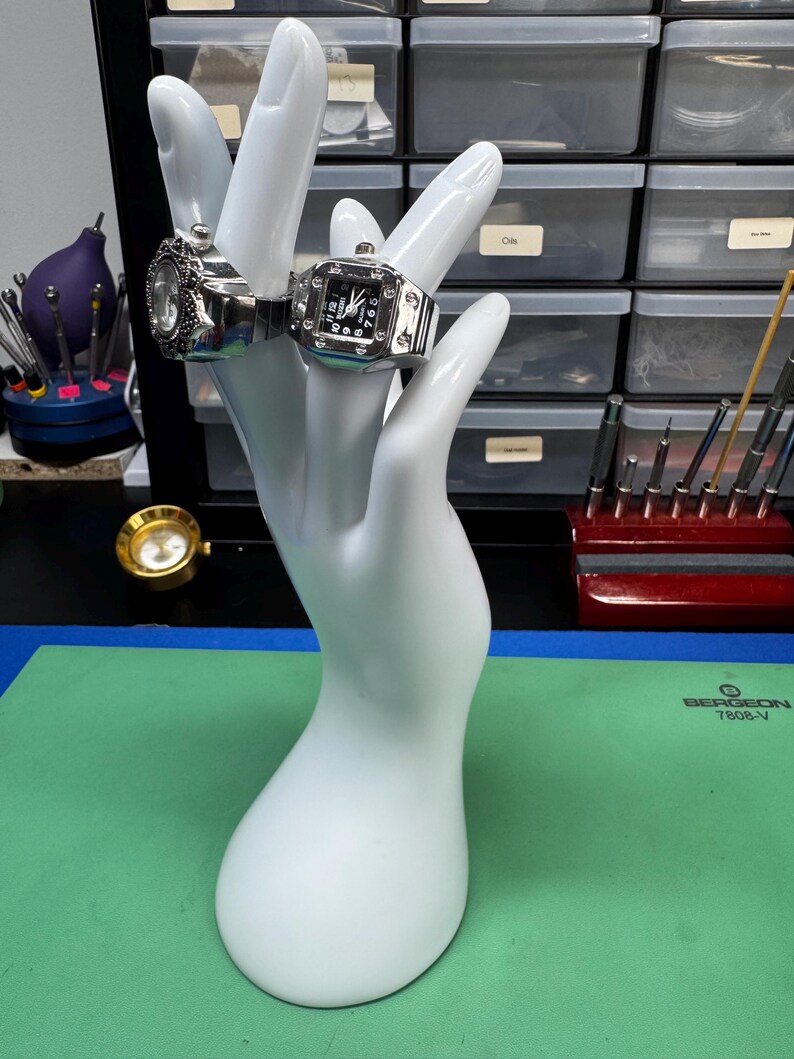 Op de afbeelding: Twee zilverkleurige horlogeringen tentoongesteld op een witte handvormige sieradenhouder. E&eacute;n ring heeft een ronde wijzerplaat, de andere een vierkante wijzerplaat met zwarte cijfers. De ringen staan tegen een achtergrond van gereedschap en opbergbakken.