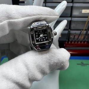 Op de afbeelding: Een zilverkleurig ringhorloge met een zwarte wijzerplaat en witte cijfers. De wijzerplaat toont de merknaam "BOZHI" en het woord "QUARTZ". Het ringhorloge wordt vastgehouden door een witte handvormige display.