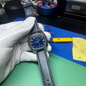 May include: A vintage wristwatch with a dark blue face and a matching blue leather strap. The watch has a rectangular silver case and a buckle closure. The watch is being held by a gloved hand.
