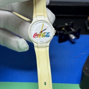 May include: A vintage white watch with a cream-colored band. The watch face features the Coca-Cola logo in red, white, and yellow. The watch hands are yellow and red. The watch is marked "SWISS".
