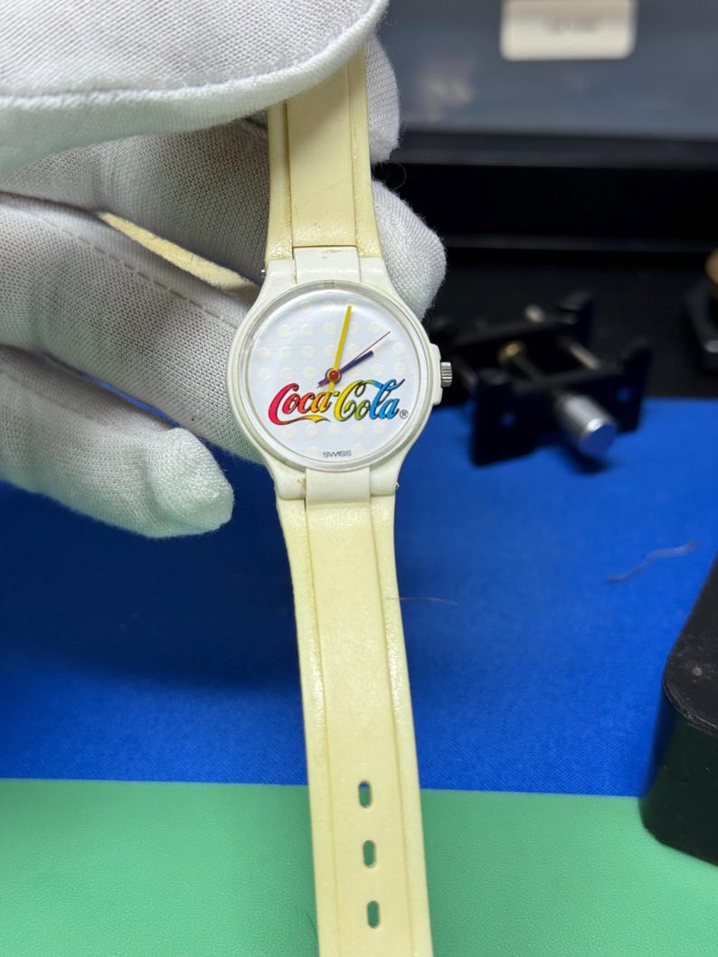May include: A vintage white watch with a cream-colored band. The watch face features the Coca-Cola logo in red, white, and yellow. The watch hands are yellow and red. The watch is marked "SWISS".