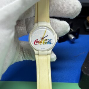 May include: A vintage Coca-Cola wristwatch with a cream-colored band and a white face featuring the Coca-Cola logo in red, yellow, and white. The watch has black hands and a yellow second hand. The watch is held by a gloved hand.