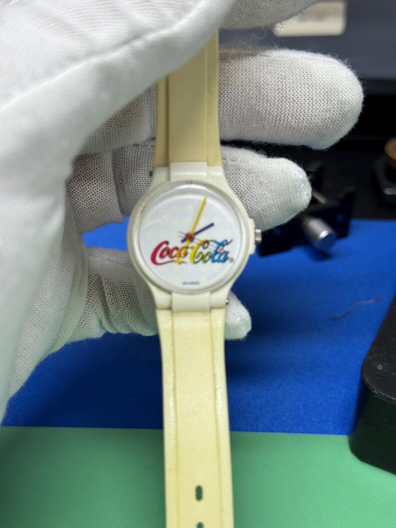 May include: A vintage Coca-Cola wristwatch with a cream-colored band and a white face featuring the Coca-Cola logo in red, yellow, and white. The watch has black hands and a yellow second hand. The watch is held by a gloved hand.