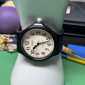 Peut inclure: Une montre Casio quartz noire avec un cadran blanc, des chiffres noirs et un bracelet noir. La montre est pr&eacute;sent&eacute;e sur une main de mannequin blanche. Le cadran porte les mots "CASIO QUARTZ" et "WATER RESIST 50M".