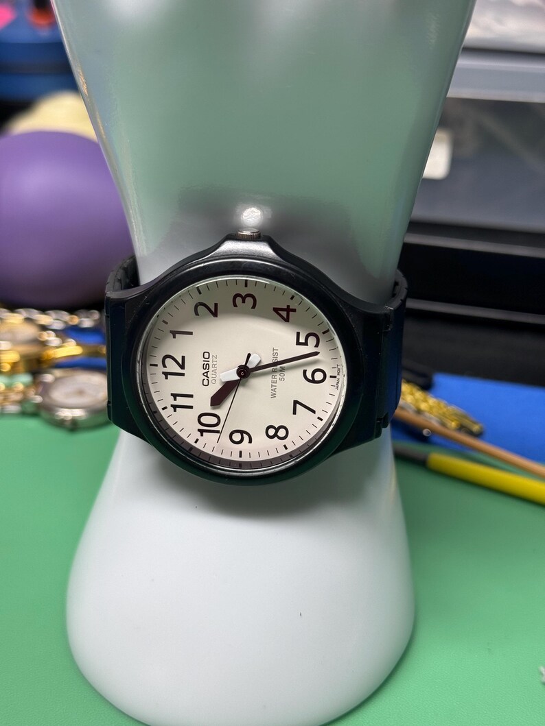 Peut inclure: Une montre Casio quartz noire avec un cadran blanc, des chiffres noirs et un bracelet noir. La montre est pr&eacute;sent&eacute;e sur une main de mannequin blanche. Le cadran porte les mots "CASIO QUARTZ" et "WATER RESIST 50M".