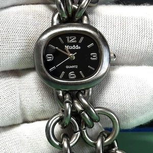 Puede incluir: Un reloj de cuarzo Mudd plateado con esfera negra y correa de cadena. La esfera muestra los números 12, 3, 6 y 9. La palabra "Quartz" está impresa debajo de las manecillas. La correa está hecha de eslabones metálicos grandes e interconectados.
