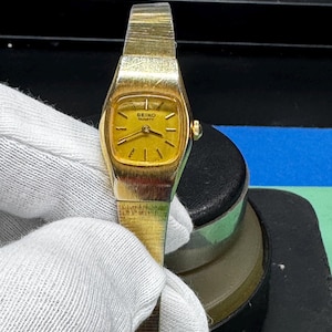 May include: A vintage gold-toned Seiko Quartz watch. The watch has a rectangular face with a gold dial and gold hands. The watch band is also gold-toned and appears to be made of metal. The watch is being held by a person wearing a white glove.