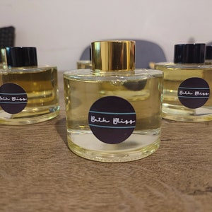Puede incluir: Cuatro botellas de vidrio transparente con tapas doradas y etiquetas negras que dicen "Bath Bliss". Las botellas están llenas de un líquido amarillo claro y están sobre una superficie de madera.