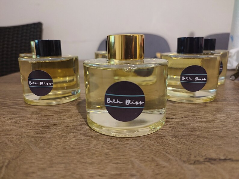 May include: Four clear glass bottles with gold lids and black labels that say "Bath Bliss". The bottles are filled with a light yellow liquid and are sitting on a wooden surface.