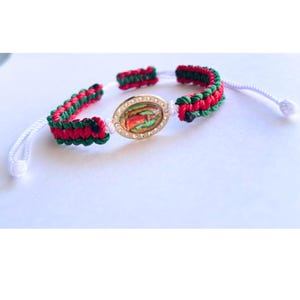 May include: A red, green, and white braided bracelet with a gold oval charm featuring a colorful image and rhinestones.