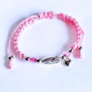 May include: Pink and white braided cord bracelet with a silver charm of the Virgin Mary, a heart charm, and pink crystal beads.