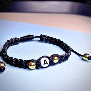 May include: A black braided bracelet with gold beads and a white bead with the letter "A" on it.
