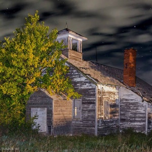 May include: A weathered, abandoned wooden building with a brick chimney and a small tower. The building is surrounded by tall grass and a large tree with yellow leaves. The sky is dark and cloudy with stars visible.