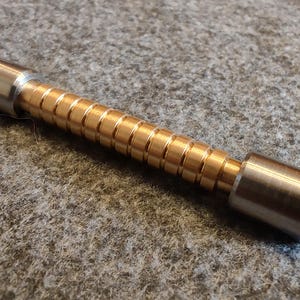 May include: A cylindrical metal object with a brass-colored, ridged center section and silver-colored ends. The object is lying on a textured, gray surface. The ridges are evenly spaced.
