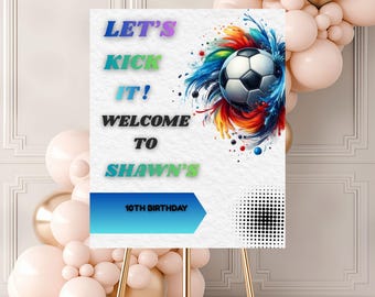 May include: A welcome sign for a 10th birthday party. The sign features the text "Let's Kick It! Welcome to Shawn's" in colorful lettering, with a soccer ball graphic and a blue arrow. The sign is on a gold easel.