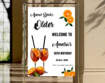 May include: A gold metal framed sign with the text "Aperol Spritz Older Welcome to Amelia's 30th Birthday 02 May, 2030". The sign features illustrations of Aperol Spritz cocktails and orange slices.