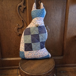 May include: A handmade cat-shaped pillow with a patchwork design. The pillow features a blue and white star pattern on the head, a patchwork of blue, black and white checkered squares, and a floral pattern on the body. A small wooden button is attached.