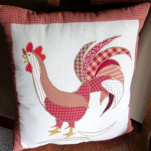 May include: A white pillow with a red, white, and brown rooster design. The rooster is made of patchwork fabric and has a large, red comb.