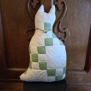 May include: A quilted cat-shaped decorative pillow in white and green. The pillow features a patchwork design with green squares and white floral patterns. A small gold bell hangs from a twine collar around the neck. The cat is sitting on a black surface.