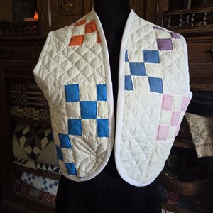 May include: A quilted, off-white vest with a diamond pattern and colorful square accents. The vest features orange, blue, and pink squares, and is displayed on a black mannequin. The edges are trimmed with white fabric.