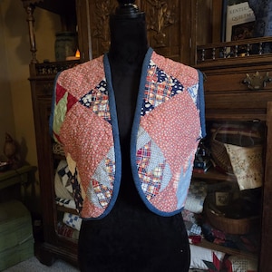 May include: A quilted vest with a patchwork design. The vest features a variety of fabrics in red, blue, and white, with a blue denim trim. The vest is displayed on a black mannequin.