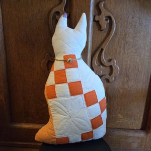 May include: A handmade cat-shaped pillow with a quilted design. The pillow features a white body with orange checkered squares and a decorative cord around the neck. The cat has pointed ears and a tail. The pillow is approximately 18 inches tall.