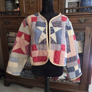 May include: A long-sleeve open-front jacket constructed from a patchwork quilt featuring a star pattern. The garment is composed of various fabric squares in shades of red, navy blue, and cream, finished with a solid cream-colored fabric trim.